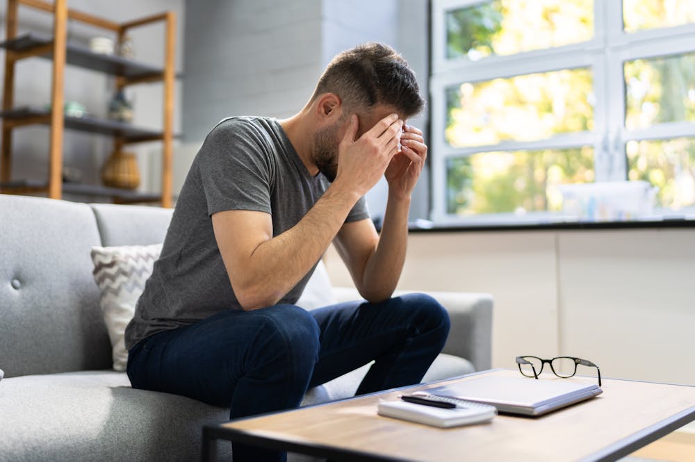Man sits on a couch, holding his head in stress, with glasses and a notebook on a table nearby.