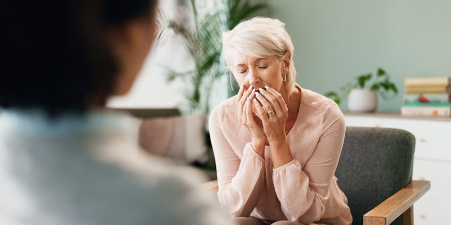 An older woman sits with closed eyes, hands covering her mouth, possibly emotional or in deep thought. She's facing another person in a calm, indoor setting.