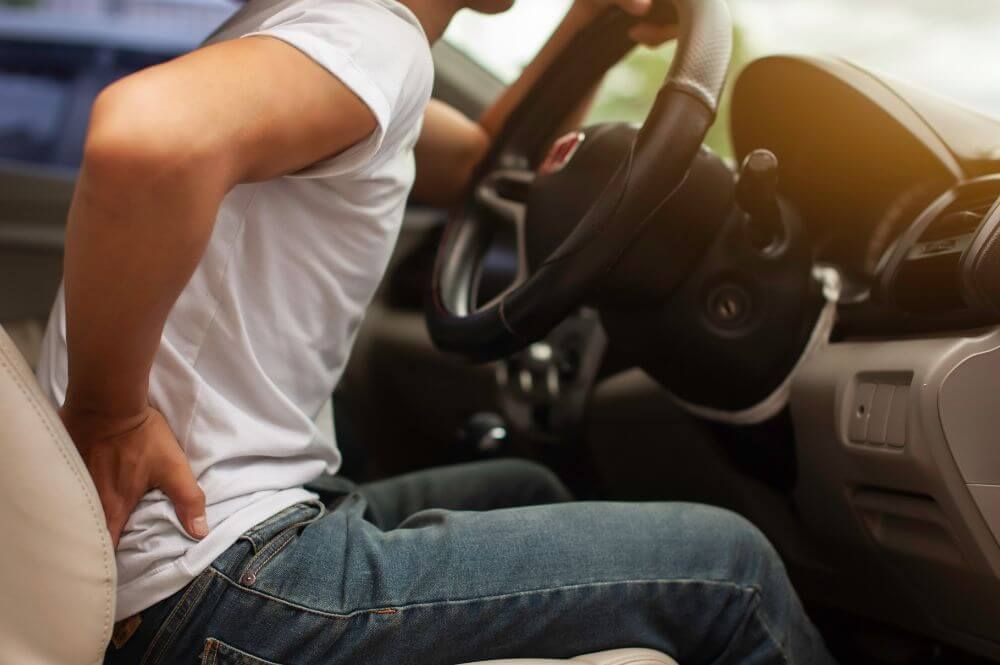A person in a white shirt sits in a car, gripping the steering wheel while wincing and holding their lower back, suggesting discomfort in the vehicle's interior.