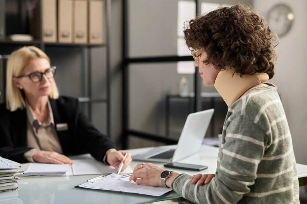 Two people sit at a desk, one signing a document; in an office with shelves and a laptop, suggesting a professional setting.