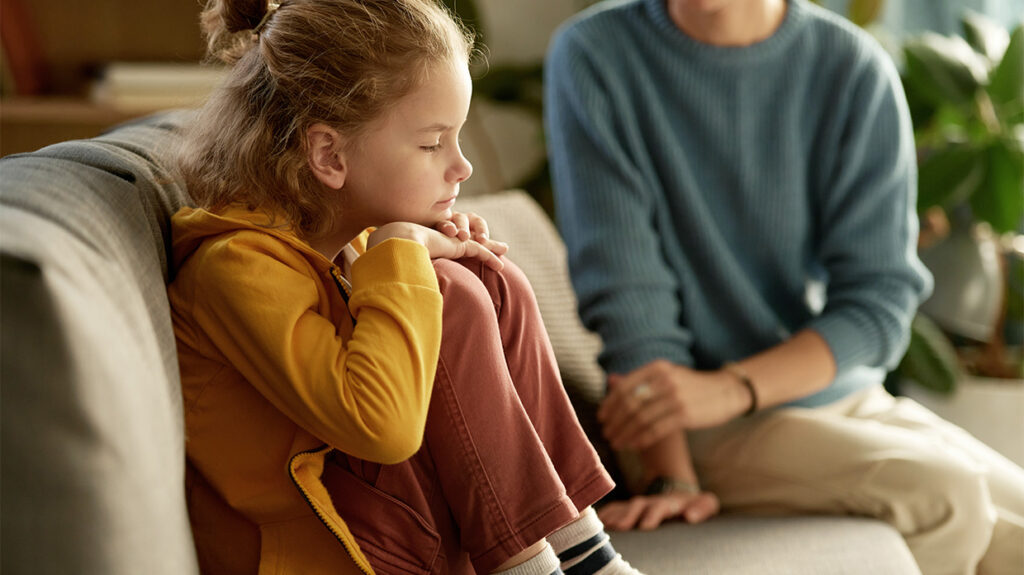 Child in a yellow jacket sitting on a couch beside an adult, looking thoughtful or concerned.