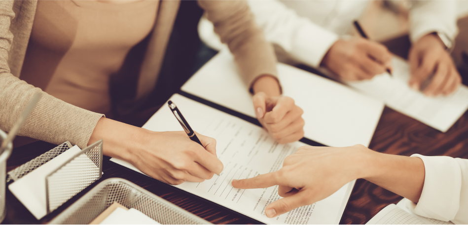 Hands sign a document, with one hand pointing at it, on a table with scattered papers and a pen. Other people are writing in the background.